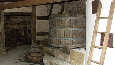 FFM_Kraisdorf_Brauerei Fotoaufnahme des Inneren des Hofbrauhauses aus Kraisdorf. Im vorderen Bereich befindet sich ein hoher Kessel mit Steinwand, an dem eine Holzleiter lehnt. Dahinter stehen ein hölzerner Sudkessel und ein Läuterbottich. Die Tür an der hintern Wand führt in den angrenzenden Raum.