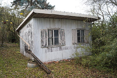 Das Haus ist klein und aus Holz. Es hat ein Pultdach. Vorne sind zwei Fenster.