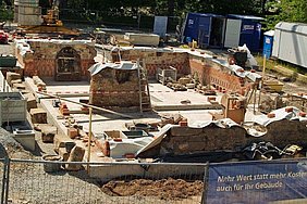 Blickrichtung Süden Fotoaufnahme der Baugrube von einem erhöhten Punkt aus. Im hinteren Teil des Erdgeschosses stehen bereits Mauern, links lehnt eine alte Holztür. In der Mitte steht ebenfalls ein Wandabschnitt, eine Leiter lehnt daran. Die Oberflächen der Wandabschnitte wurden mit Planen verdeckt.