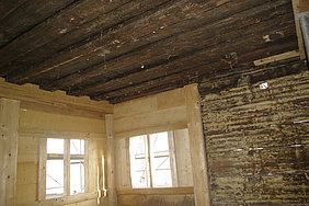 Bohlen-Balkendecke aus dunklem Holz oberhalb einer vertäfelten Stube