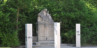 FFM_Kriegerdenkmal_Ornbau ein Steindenkmal mit drei Figuren - eine "Pietas" mit sterbendem Krieger auf dem Schoß, daneben ein "Jüngling", dem ein Schwert gereicht wird. Drei Informationsstelen kontextualisieren das Denkmal.