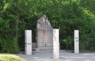 FFM_Kriegerdenkmal_Ornbau ein Steindenkmal mit drei Figuren - eine "Pietas" mit sterbendem Krieger auf dem Schoß, daneben ein "Jüngling", dem ein Schwert gereicht wird. Drei Informationsstelen kontextualisieren das Denkmal.