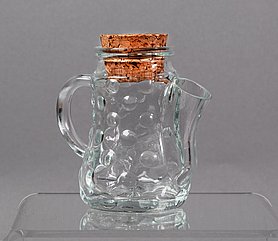 Die Kanne ist aus Glas. Sie hat runde Noppen. An einer Seite ist ein Henkel. An der anderen Seite ist ein Ausguss. Oben ist eine Öffnung. In der Öffnung steckt ein Korken.