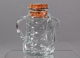 Die Kanne ist aus Glas. Sie hat runde Noppen. An einer Seite ist ein Henkel. An der anderen Seite ist ein Ausguss. Oben ist eine Öffnung. In der Öffnung steckt ein Korken.