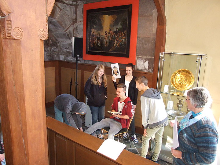 Impressionen aus der Aktion "Und woher kommst du?" Fotoaufnahme der Aktion "Und woher kommst du?" in der Spitalkirche. Im Fokus steht eine Gruppe aus fünf Jugendlichen und einer Erwachsenen. Mittig sitzt ein Junge mit Buch auf einem Stuhl, das Mädchen dahinter hält ein Bild hoch. Im Hintergrund hängt ein Gemälde der Taufe Jesu.