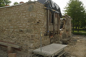 Ergänzte Teilabschnitte Fotoaufnahme der ergänzten Teilabschnitte. Im Fokus steht eine Ecke des Erdgeschosses. Auf der Oberseite des Bruchsteinmauerwerks liegt eine Plane, daneben steht eine heruntergefahrene Hebebühne. Die Mauer auf der rechten Seite ist noch nicht vollständig.