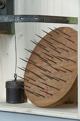 Die runde Scheibe ist aus Holz. Auf ihr sind sehr viele Nagelspitzen. Auf die Spitzen wurden Pinselstiele gesteckt.