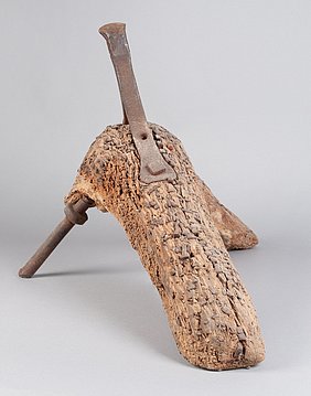 Der Beschlagbock ist aus Holz. Das Holz ist sehr grob. In dem Holz stecken viele Nägel. Auf dem Bock ist eine Leiste aus Eisen.