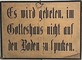 Das Schild ist hellbraun. Es hat einen schwarzen Rahmen. Auf dem Schild steht: Es wird gebeten, im Gotteshaus nicht auf den Boden zu spucken.