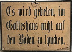 Das Schild ist hellbraun. Es hat einen schwarzen Rahmen. Auf dem Schild steht: Es wird gebeten, im Gotteshaus nicht auf den Boden zu spucken.