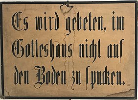 Das Schild ist hellbraun. Es hat einen schwarzen Rahmen. Auf dem Schild steht: Es wird gebeten, im Gotteshaus nicht auf den Boden zu spucken.