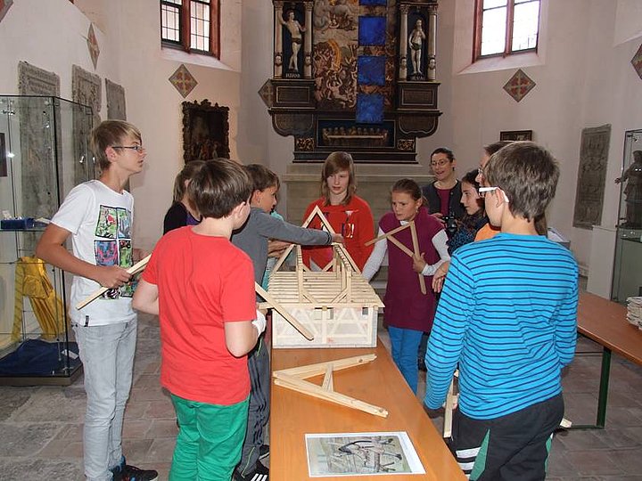 FFM_Unter_dem_Dach_der_Kirche“ Fotoaufnahme der Führung „Unter dem Dach der Kirche“ in der Bad Windsheimer Spitalkirche. Eine Gruppe Kinder steht um einen Bierzelttisch. Darauf befindet sich ein Haus-Modell, anhand dessen sie ein Dach konstruieren. Im Hintergrund ist ein Altar zwischen zwei Fenstern der Kirche erkennbar.