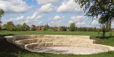 Fotoaufnahme des grünen Klassenzimmers auf dem Erlebnisplatz in der Baugruppe Süd.  Aus Steinen wurde eine Rotunde gefertigt. In einem Halbkreis sind drei Sitzreihen stufenartig übereinander angeordnet. Im Hintergrund sind die Gebäude des Museums erkennbar. 