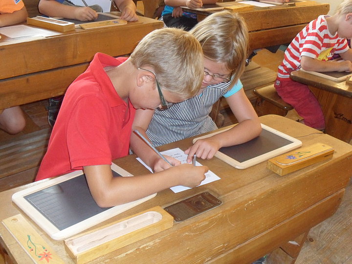 Fotoaufnahme des Kinderferienprogramms. An alten Schulbänken sitzen je zwei Kinder, die mit Griffeln auf Schiefertafeln schreiben. Die Tafeln der beiden vorderen Kinder sind noch leer, sie üben das Griffelschreiben auf einen Blatt Papier, das zwischen ihnen liegt. 
