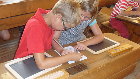 FFM_Kinderferienprogramm Fotoaufnahme des Kinderferienprogramms. An alten Schulbänken sitzen je zwei Kinder, die mit Griffeln auf Schiefertafeln schreiben. Die Tafeln der beiden vorderen Kinder sind noch leer, sie üben das Griffelschreiben auf einen Blatt Papier, das zwischen ihnen liegt.