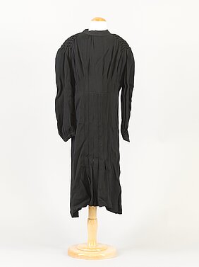 Das Kleid ist schwarz. Es hat lange Ärmel. Der Stoff ist faltig.