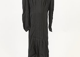 Das Kleid ist schwarz. Es hat lange Ärmel. Der Stoff ist faltig.
