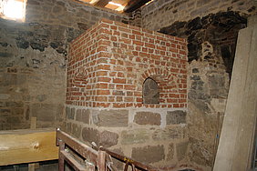 28.8.18 Schwitzofen. Der fertige Bau zeigt die Größe und Dominanz des Schwitzofens innerhalb der Badstube. Fotoaufnahme des fertigen Schwitzofens in einer Ecke der Badstube. Der untere Teil besteht aus Sandsteinquadern, der obere wurde mit Ziegelsteinen rekonstruiert. An den beiden sichtbaren Seiten ist je eine rundbogige Öffnung im Ziegelmauerwerk erkennbar.