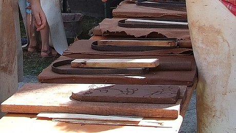 FFM_Ziegeln Fotoaufnahme der Handwerkervorführung. Im Vordergrund steht eine Bierzeltgarnitur mit Ziegelformen und einem fertigen Feierabendziegel. Eine Frau in Schütze steht dahinter und erklärt zwei Besuchern gerade etwas. Im Hintergrund steht ein weiterer Tisch, an dem Ofenkacheln geformt werden.