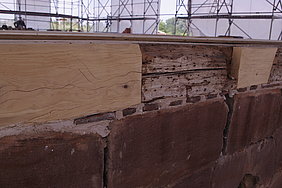 6.6.18 Nach ihrer endgültigen Ausrichtung waren die auf der Mauer liegenden Hölzer noch mit Mörtel, Bruchsteinen oder Ziegelresten zu untermauern. Fotoaufnahme der auf dem Sandsteinmauerwerk liegenden Holzbalken. Diese wurden mit Mörtel, Bruchsteinen und Ziegelresten untermauert. Der Holzbalken wurde an zwei Stellen mit frischem Nadelholz repariert. Im Hintergrund ist das teilweise mit Planen verhangene Gerüst erkennbar.