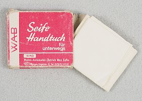 Die Schachtel ist rechteckig. Sie ist rot bedruckt. Die Schrift ist weiß. Das weiße Handtuch liegt daneben. Es ist gefaltet.