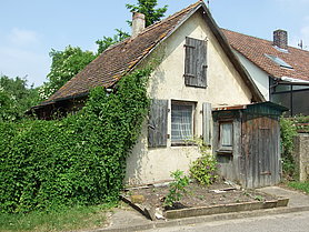 FFM_Aussen Fotoaufnahme des Behelfsheimes aus Ottenhofen am alten Standort. Vor dem kleinen Haus befindet sich ein Beet, in dem nur wenige Pflanzen stehen. An der Hauswand wächst links eine Kletterpflanze. Im Hintergrund sind links Bäume erkennbar, rechts steht das Nachbarhaus.