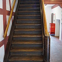 FFM_Gasthaus_zur_Krone_Treppenaufgang Fotoaufnahme des Treppenaufgangs im Gasthaus zur Krone aus Oberampfrach. Die 17-stufige Holztreppe mit dem Geländer auf beiden Seiten führt in das Obergeschoss. Am Fuß der Treppe steht ein Stuhl aus Holz, die Wände und die Decke sind im Fachwerkstil gehalten.