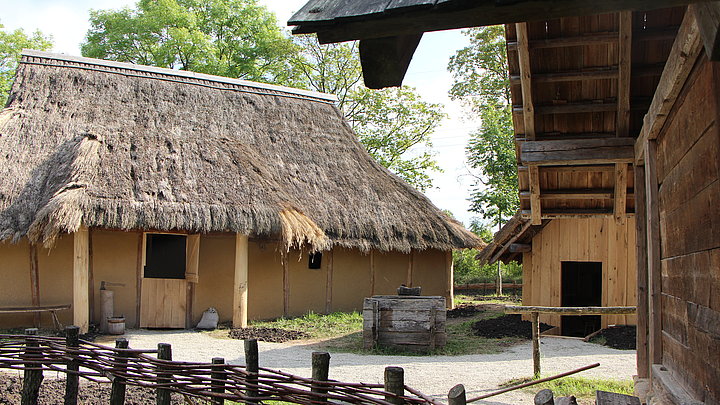 FFM_Eroeffnung_Archaeologisches_Dorf Fotoaufnahme des Archäologischen Dorfes. Im Vordergrund steht der mit Weiden durchflochtene Zaun. Dahinter liegt links das rekonstruierte Wohnhaus, ein Firstpfostenhaus mit Strohdach. Rechts daneben befinden sich die Werkstatt und der Speicher. In der Mitte steht ein Brunnen.