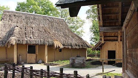 FFM_Eroeffnung_Archaeologisches_Dorf Fotoaufnahme des Archäologischen Dorfes. Im Vordergrund steht der mit Weiden durchflochtene Zaun. Dahinter liegt links das rekonstruierte Wohnhaus, ein Firstpfostenhaus mit Strohdach. Rechts daneben befinden sich die Werkstatt und der Speicher. In der Mitte steht ein Brunnen.