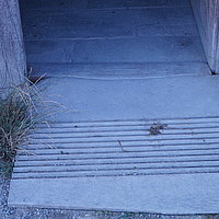 FFM_West_Schaeferei_Rampe_5 Fotoaufnahme der Schwelle am rechten Eingang der Schäferei aus Hambühl am aktuellen Standort. Vor der Schwelle liegt eine Rampe aus Holz, links und rechts davon wächst Gras. Der Boden im Inneren ist mit Steinplatten gepflastert.