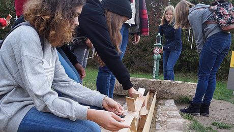 FFM_Wasserraedchen Ein paar Schülerinnen testen ihre selbstgebauten Wasserrädchen in einer Holzrinne an einem Pumpbrunnen