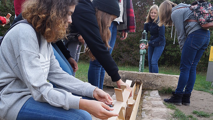 FFM_Wasserraedchen Ein paar Schülerinnen testen ihre selbstgebauten Wasserrädchen in einer Holzrinne an einem Pumpbrunnen