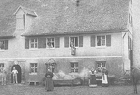 Vor einem alten Haus stehen fünf Personen, zwei Männer, drei Frauen; aus drei der geöffneten Fenster des 1. Stockwerks schaut je eine Person heraus, zwei Männer, eine Frau; Alle Personen sind dem Fotografen zugewandt und schauen in die Kamera
