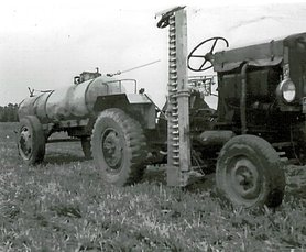 Der Schlepper zieht ein Wasserfass. Er hat ein Mähwerk, mit dem man Gras mähen kann.