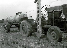 Der Schlepper zieht ein Wasserfass. Er hat ein Mähwerk, mit dem man Gras mähen kann.