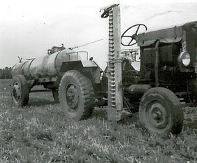 Der Schlepper zieht ein Wasserfass. Er hat ein Mähwerk, mit dem man Gras mähen kann.