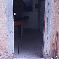 FFM_West_Oelmuehle_Muehlraum_Eingang Fotoaufnahme des vorderen Eingangs zum Mühlraum der Ölmühle von der Fledrichsmühle am aktuellen Standort. Vor der Türschwelle liegt eine Rampe aus Holz, der Boden im Inneren besteht aus flachen Steinplatten. Neben der Tür blühen Wildblumen.