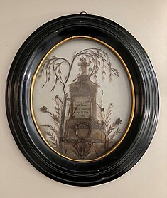 Das Bild ist oval. Der Rahmen ist aus Holz. Das Bild zeigt ein Denkmal. Es besteht aus Zweigen und Haaren.