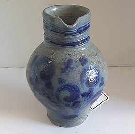 Die Kanne ist rundlich. Sie ist blau bemalt. Sie hat einen Ausguss.