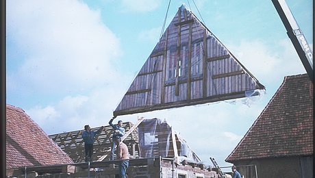 FFM_Giebel_am_Haken Fotoaufnahme eines verschalten Giebels am Haken eines Krans. Ein Giebel wird gerade auf das Steinmauerwerk des Ergersheimer Häckerhaus gesetzt. An der Mauer steht ein Gerüst, drei Männer stehen darauf bzw. auf der Mauer, um bei der Positionierung zu helfen.