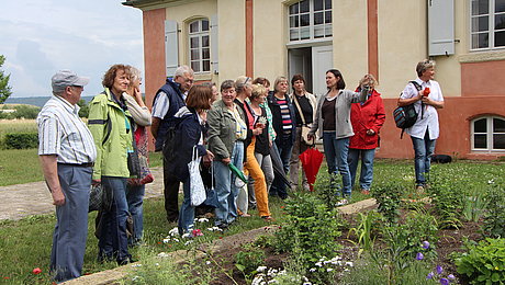 Fotoaufnahme einer Führung im Garten des Jagdschlösschens aus Eyerlohe. Die Gruppe steht vor dem angelegten Blumenbeet. Es blühen unter anderen Staudenpfingstrosen, Mohn und Clematis. Hinter der Gruppe befindet sich das Schlösschen mit der geöffneten Tür. 