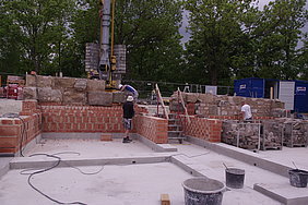 Fotoaufnahme des Aufbaus der ersten historischen Mauerabschnitte. Auf die Mauer aus Hochlochziegeln werden die historischen Sandsteinquader mit Hilfe eines Karns aufgesetzt. Mittig befindet sich eine Bautreppe. Vor der Baumreihe und dem Bauzaun im Hintergrund steht der Kran. 