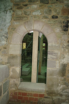 Türabschluss, Ansicht Richtung Nord vom Flur Fotoaufnahme einer fertiggestellten Tür im Badhaus. Das Foto wurde im Flur aufgenommen. Gezeigt wird der runde Torbogen aus Sandsteinquadern, eingebettet in das Steinmauerwerk. Im Raum dahinter sind aufgestellte Holzbalken und Metallstreben erkennbar.