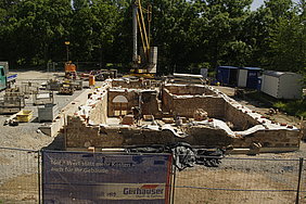 Fotoaufnahme der Baugrube von einem erhöhten Standpunkt. Die Grundmauern stehen bereits, die Oberflächen der Bauteile wurden mit Planen verdeckt. In der Baugrube liegen Werkzeug, Steine und andere Materialien, einige Männer sind bei der Arbeit. Dahinter steht ein Kran. 