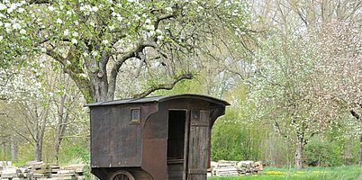 Fotoaufnahme des Wagens auf dem Erlebnisplatz in der Baugruppe Süd. Der überdachte Einachser erinnert mit seinem kleinen vergitterten Fenster und der Holztür stark an einen Gefängniswagen. Im Hintergrund stehen blühende Bäume. 