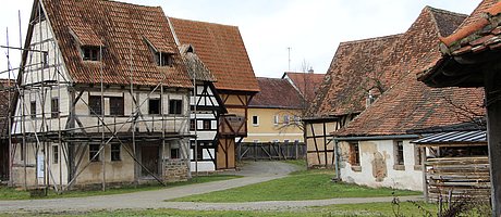 Fotoaufnahme der Gasse mittelalterlicher Fachwerkhäuser in der Baugruppe Stadt. An einem Weg befinden sich auf beiden Seiten verschiedene Fachwerkhäuser. Vorne links steht zum Beispiel das Handwerkerhaus aus Bad Windsheim, das von einem Holzgerüst umgeben ist.  