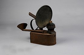Die Lampe hat einen Behälter. Auf dem Behälter ist ein Schirm. An der Lampe ist ein Tragegriff.