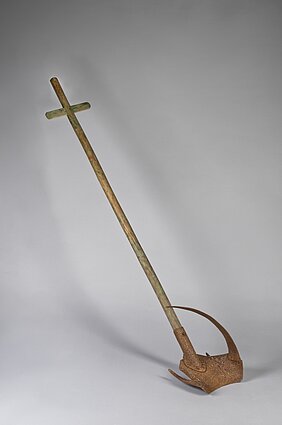 Das Gerät hat einen Holzstiel. Am Stiel ist eine Schippe. Die Schippe hat einen Metallbogen. Unten an der Schippe sind Kufen.