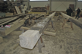 FFM_Bautagebuch_Wendelstein_Balkenlage Fotoaufnahme der Reparaturen an den Erdgeschossdeckenbalken in der Abbundhalle der Firma Lederer. Im Vordergrund liegen zwei Balken auf Holzböcken. Die Schäden daran wurden bereits mit frischem Nadelholz repariert. Links dahinter liegen weitere Balken, rechts stehen drei Männer.