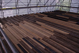 Fotoaufnahme der Erdgeschossbalkenlage auf ihrer endgültigen Position. Das Foto wurde von einem erhöhten Standpunkt aufgenommen. Am Großteil der Holzbalken wurden Reparaturen mit Nadelholz oder Stahlträger vorgenommen. Dahinter ist das mit Planen verhangene Gerüst erkennbar. 